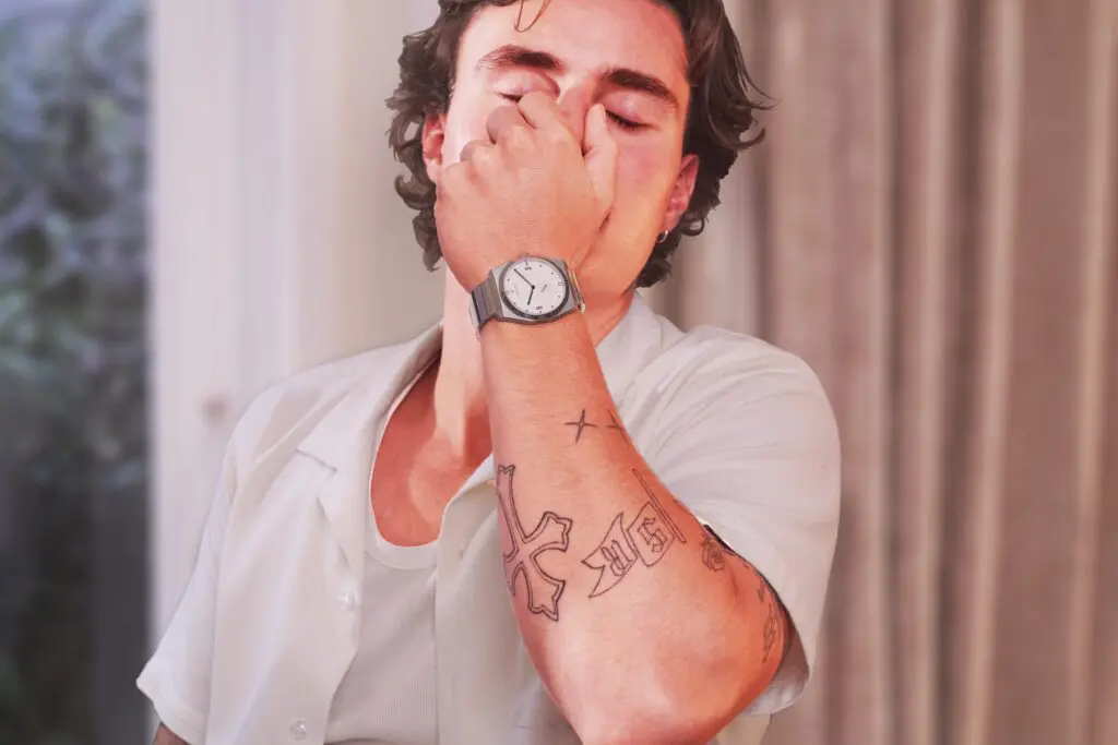 A man in a white shirt covers his face with his hand, a Komposition Cream on his wrist
