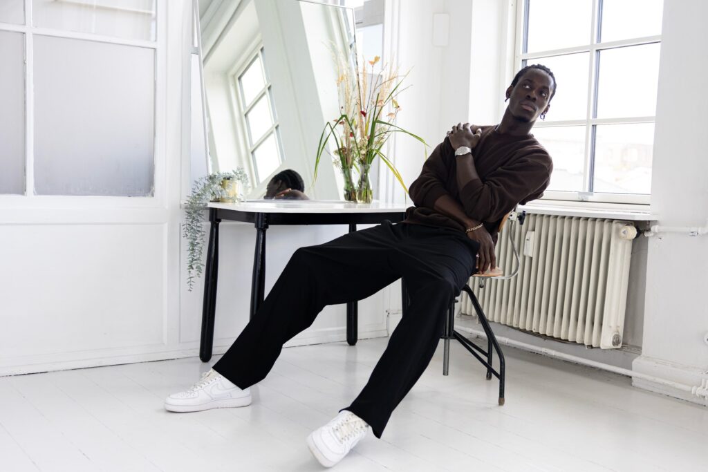 A man leans back on a chair by a bright window, a Komposition Automatic on his wrist